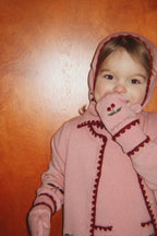 Cagoule en laine Collette – Pale Blush – Konges Sløjd : enfant avec un gilet rose à cerises tenant sa manche près du visage.