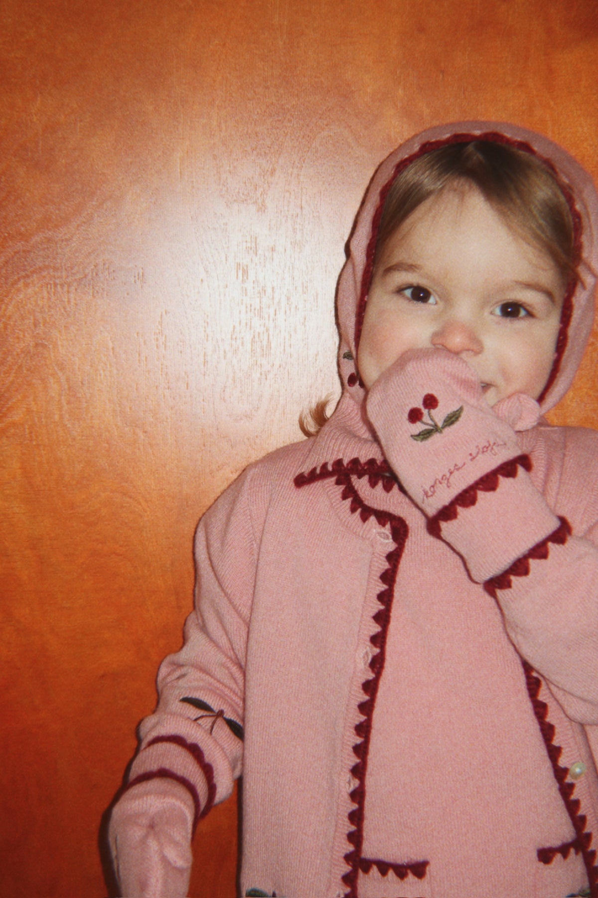 Cagoule en laine Collette – Pale Blush – Konges Sløjd : enfant avec un gilet rose à cerises tenant sa manche près du visage.