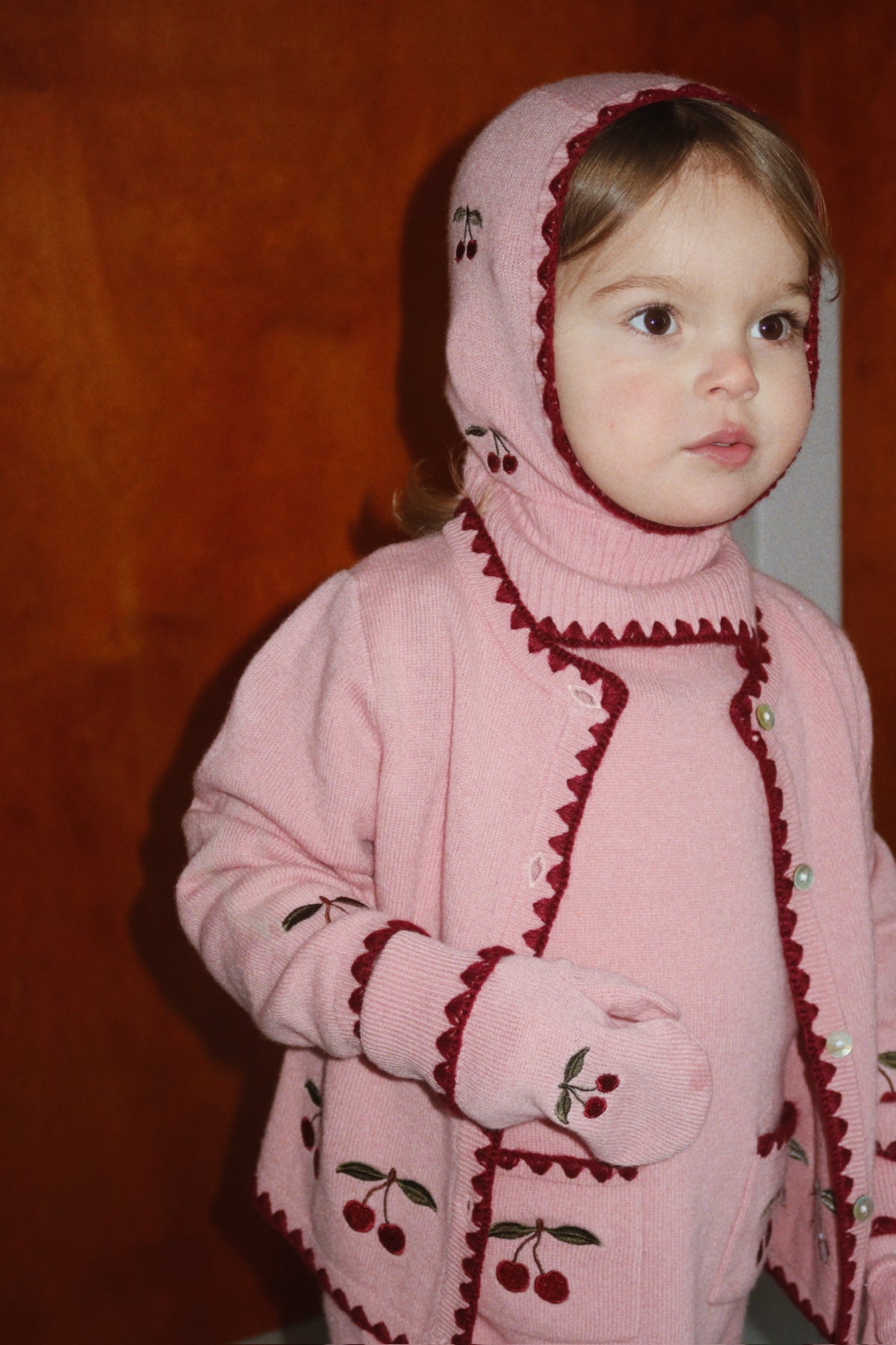 Cagoule en laine Collette – Pale Blush – Konges Sløjd : enfant portant un gilet rose et la cagoule à motif cerise assortie.